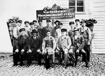 Eine Gruppe von Männern in historischen Arbeitskleidern steht vor einem Schild mit der Aufschrift „Wachonheimer Wasserzubehörgesellschaft“. Die Aufnahme scheint aus einer vergangenen Zeit zu stammen und zeigt eine Verbindung zur lokalen Industrie.