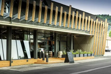 Ein modernes Gebäude mit Holz- und Glaselementen. Vor dem Eingang stehen Pflanzen und Fässer, die eine einladende Atmosphäre schaffen.