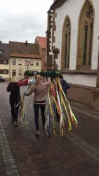 Zwei Personen tragen einen bunten Maibaum mit vielen Bändern auf einer Straße. Im Hintergrund ist eine Kirche zu sehen.