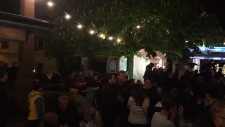 Eine belebte Veranstaltung im Freien bei Nacht, mit vielen Menschen und Lichtern. Es gibt Bäume und Stände, die eine festliche Stimmung schaffen.