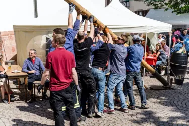 Eine Gruppe von Männern hebt gemeinsam einen langen Balken an. Im Hintergrund sind Tische und andere Personen bei einer festlichen Veranstaltung zu sehen.