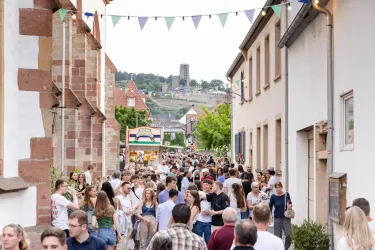 Eine lebhafte Straße voller Menschen mit bunten Wimpeln im Hintergrund. Im Hintergrund sind Gebäude und eine Hügelkulisse zu sehen.