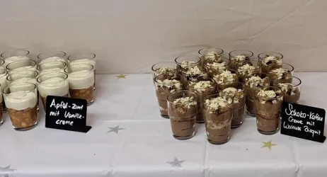 Auf dem Tisch stehen Gläser mit zwei verschiedenen Dessertvariationen. Links sind Apfel-Zimt mit Vanillecreme und rechts ist Schoko-Kaffee mit einer Schicht Sahne.