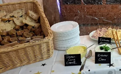 Ein Korb mit verschiedenen Brotsorten steht neben weißen Tellern. Daneben befinden sich verschiedene Dips in Schalen, beschriftet mit „Dattel-Curry-Creme“ und „Kräuter-Creme“.