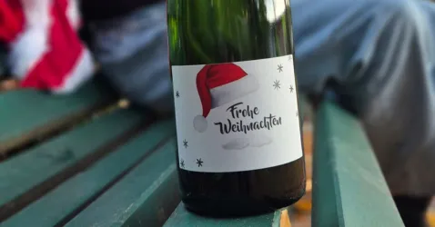 Eine Flasche Sekt mit einem Etikett, das eine Weihnachtsmütze und den Schriftzug „Frohe Weihnachten“ zeigt. Der Hintergrund zeigt eine Holzbank und eine Person in festlicher Kleidung.