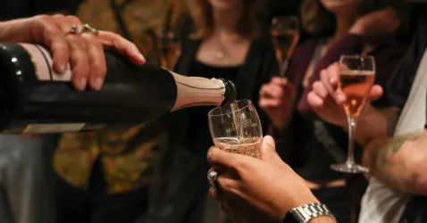 Eine Person schenkt Sekt in ein Glas ein. Im Hintergrund stehen weitere Gäste mit Gläsern.
