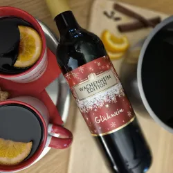 Eine Flasche Glühwein steht neben zwei roten Tassen, gefüllt mit Glühwein und dekoriert mit Orangenscheiben. Im Hintergrund sind Zimtstangen und eine Schüssel mit mehr Glühwein zu sehen.