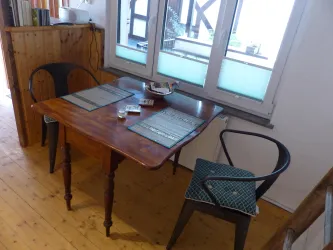 Ein gemütlicher Tisch mit zwei Stühlen in einem hellen Raum. Auf dem Tisch liegen Tischsets und ein Glas.