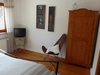 Ein gemütliches Zimmer mit einem Bett, einem Fernseher und einem Holzschrank. An der Wand hängen zwei Bilder und ein Sessel steht in der Ecke.