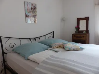 Ein gemütliches Schlafzimmer mit einem eisernen Bett und hellblauen Kissen. Im Hintergrund steht eine Holzkommode und ein Dekorbild an der Wand.