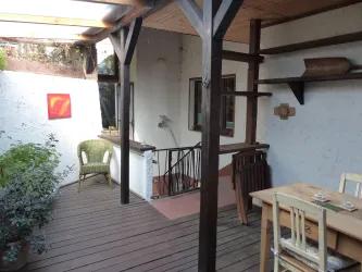 Eine gemütliche Terrasse mit Holzfußboden und einem Tisch. Im Hintergrund stehen ein Stuhl und eine Treppe in ein helles Zimmer.