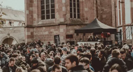 Eine große Menschenmenge versammelt sich vor einem Gebäude mit einer schwarzen Markise. Die Stimmung scheint lebhaft und gesellig zu sein.