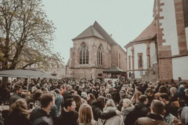 Eine große Menschenmenge versammelt sich vor einer historischen Kirche. Es herrscht eine festliche Atmosphäre mit Bäumen und Ständen im Hintergrund.