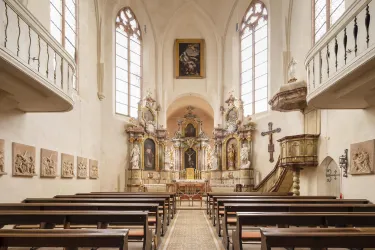Eine ruhige Kirche mit Holzbänken und hohen Fenstern. Der Altarbereich ist kunstvoll gestaltet mit Skulpturen und Bildern.
