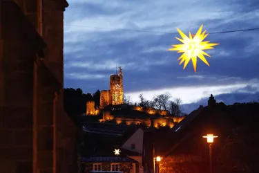 Eine beleuchtete Burg im Hintergrund, umgeben von Bäumen, leuchtet in der Dämmerung. Im Vordergrund hängt ein großer, leuchtender Stern.