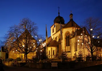 Eine Kirche bei Nacht, beleuchtet von zarten Lichtern. Im Hintergrund stehen kahlen Bäumen und ein klarer, blauer Himmel.