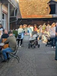 Eine belebte Außenszene mit Menschen, die an Tischen sitzen und sich unterhalten. Im Hintergrund sind Kinderwagen und eine Gruppe von Personen zu sehen, die miteinander sprechen.