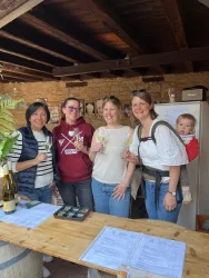 Fünf Frauen stehen an einem Tisch in einem rustikalen Raum und halten Sektglas in der Hand. Eine Frau trägt ein Baby auf dem Rücken und alle lächeln.