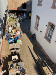 Ein gemütlicher Innenhof mit mehreren Tischen und Gästen, die sich unterhalten. Die Sonne scheint und es herrscht eine angenehme Atmosphäre.