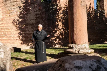 Ein Mann in einem schwarzen Gewand steht zwischen alten Steinen und einer Säule. Die Sonne wirft Schatten auf die Ruinen im Hintergrund.