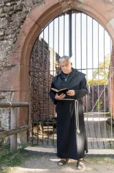 Ein Mönch steht vor einem von Gitterstäben gesicherten Tor und liest in einem Buch. Die Kulisse zeigt eine alte, steinerne Mauer und einen grünen Pfad.