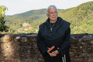 Ein älterer Mönch steht an einer Wand mit Blick auf eine grüne Hügellandschaft. Er trägt eine schwarze Kutte und hält ein Buch in der Hand.