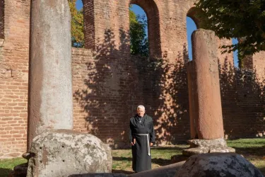 Eine Person steht zwischen alten Säulen in einer historischen Ruine. Im Hintergrund sind massive, rötliche Mauern und Bäume zu sehen.