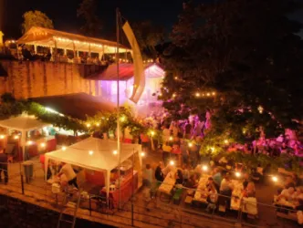 Eine belebte Outdoor-Veranstaltung bei Nacht mit bunten Lichtern und festlicher Stimmung. Menschen sitzen an Tischen und genießen die Atmosphäre.