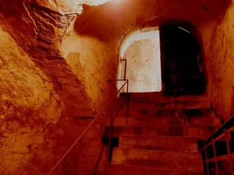 Eine düstere Treppe führt hinunter in einen alten Keller. Die Wände sind rustikal und der Lichtschein ist schwach.