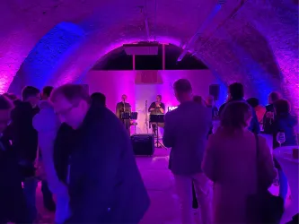 Ein lebhaftes Event in einem künstlerisch beleuchteten Raum mit einer Band auf der Bühne. Menschen unterhalten sich und genießen die Atmosphäre.
