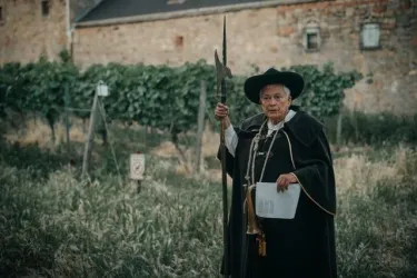Ein Mann in traditioneller Kleidung steht in einem Weinberg. Er hält eine Pflanze und scheint die Umgebung zu überwachen.