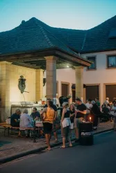 Eine lebhafte Szene vor einem historischen Gebäude mit Menschen, die an Tischen sitzen und sich unterhalten. Die Atmosphäre ist entspannt und gesellig, während es langsam dunkel wird.