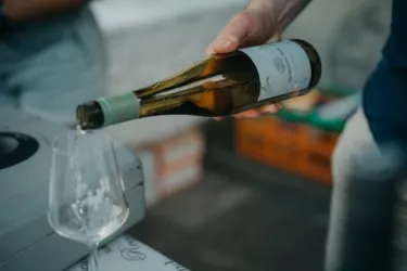 Eine Person schenkt Wein aus einer Flasche in ein Glas. Der Hintergrund zeigt eine entspannte Atmosphäre mit Kisten und einem Tisch.