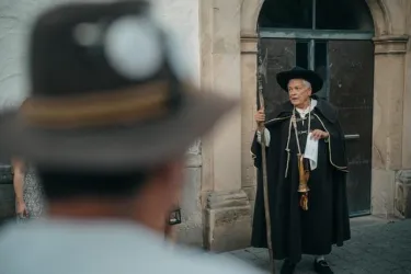 Ein Mann in traditioneller Kleidung steht mit einem Stock in der Hand. Im Hintergrund ist ein historisches Gebäude sichtbar.