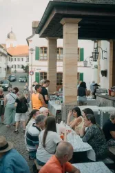 Eine belebte Freiluft-Gastronomie mit vielen Gästen. Es gibt Tische und Stühle, und die Atmosphäre ist gesellig und entspannt.