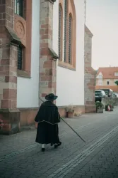 Eine Person in einem schwarzen Umhang und Hut geht entlang einer Kirche. Der Weg ist gepflastert und im Hintergrund sind einige Gebäude sichtbar.