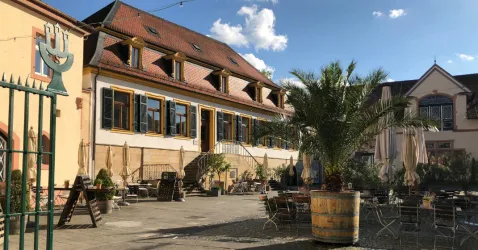 Ein einladender Außenbereich eines Restaurants mit Sitzgelegenheiten und Sonnenschirmen. Im Hintergrund ist ein historisches Gebäude mit bunten Fenstern und einem schönen Himmel zu sehen.