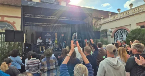 Eine Musikergruppe tritt auf einer Bühne auf, während das Publikum begeistert applaudiert. Im Hintergrund ist ein historisches Gebäude und eine sonnige Wetterlage zu sehen.