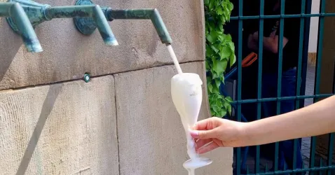 Ein Glas wird unter einem Wasserhahn gehalten, aus dem ein schaumiges Getränk fließt. Im Hintergrund sind grüne Pflanzen und ein Tor sichtbar.