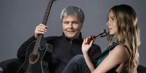 Ein Mann mit einer Gitarre sitzt auf einem Sessel und lächelt. Neben ihm sitzt eine Frau, die nachdenklich einen Pinsel hält.