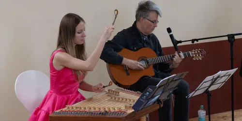 Eine Musikerin in einem pinken Kleid spielt auf einer Zither, während ein Begleiter auf der Gitarre spielt. Die Atmosphäre ist entspannt und musikalisch.
