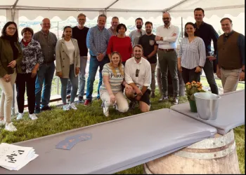 Eine Gruppe von Personen posiert gemeinsam unter einem Zelt im Freien. Im Vordergrund sind ein Tisch und eine Blumenvase sichtbar.