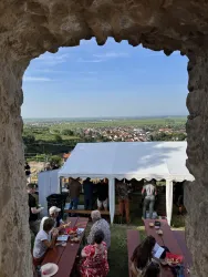 Ein Blick durch einen alten Steinbogen auf eine Veranstaltung im Freien. Im Vordergrund sind Tische und Stühle zu sehen, während im Hintergrund die Landschaft und ein Zelt mit Gästen sichtbar sind.