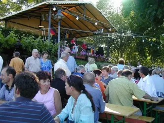 Eine lebendige Veranstaltung im Freien mit vielen Menschen, die an Tischen sitzen und sich unterhalten. Im Hintergrund gibt es eine Bühne mit Musikern und festlicher Beleuchtung.
