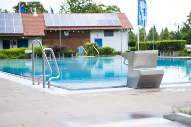 Ein klarer Pool steht vor einem Gebäude mit Solarzellen auf dem Dach. Die Umgebung ist gepflegt und mit grünen Pflanzen ausgestattet.