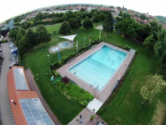 Ein Schwimmbecken in einer grünen Umgebung mit Bäumen und Wiese. Im Hintergrund sind Wohngebäude und ein Parkplatz zu sehen.