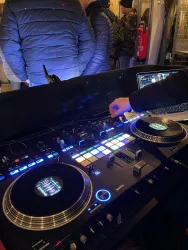 Ein DJ-Mischpult mit Turntables und einem Laptop. Im Hintergrund sind Menschen zu sehen, die zuhören.