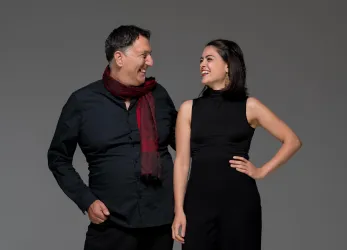 Ein Mann und eine Frau stehen nebeneinander und lachen. Beide tragen schwarze Kleidung, der Mann hat einen roten Schal um den Hals.