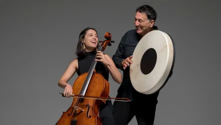 Ein Künstlerehepaar spielt gemeinsam Musik. Die Cellistin lächelt, während der andere Musiker mit einem Schlaginstrument begleitet.