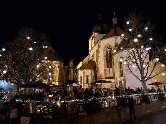 Ein belebter Platz bei Nacht mit festlicher Beleuchtung und einer Kirche im Hintergrund. Viele Menschen genießen die Atmosphäre unter den leuchtenden Sternen.